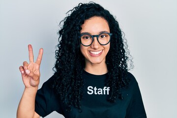 Young hispanic woman with curly hair wearing staff t shirt smiling looking to the camera showing fingers doing victory sign. number two.