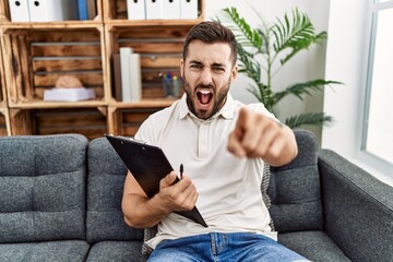 Handsome hispanic man holding clipboard working at psychology clinic pointing displeased and frustrated to the camera, angry and furious with you