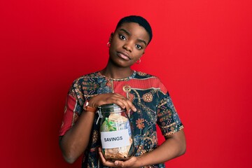 Young african american woman holding savings jar with south african rands money relaxed with serious expression on face. simple and natural looking at the camera.