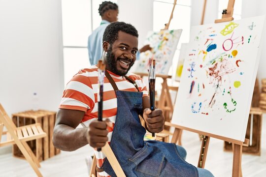 African american painter couple smiling happy painting at art studio. Man holding paintbrushes sitting on chair.