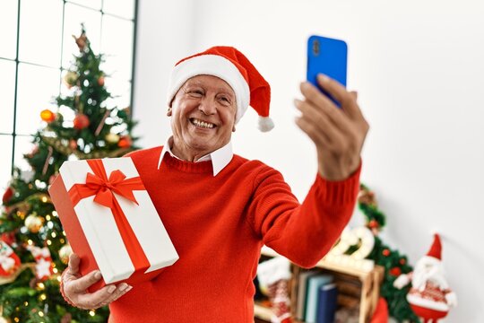 Senior grey-haired man holding christmas gift making selfie by the smartphone at home.