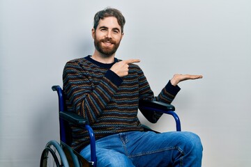 Caucasian man with beard sitting on wheelchair amazed and smiling to the camera while presenting...