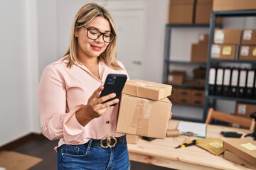 Young hispanic woman ecommerce business worker holding packages and using smartphone at office