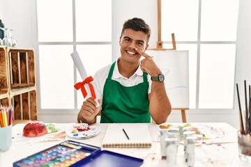 Young hispanic man at art studio holding degree pointing with hand finger to face and nose, smiling cheerful. beauty concept