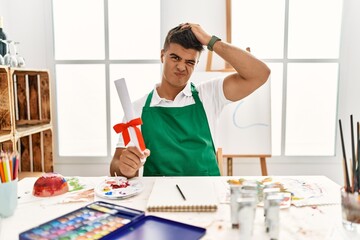Young hispanic man at art studio holding degree confuse and wondering about question. uncertain with doubt, thinking with hand on head. pensive concept.