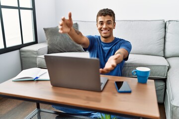 Young handsome hispanic man using laptop sitting on the floor looking at the camera smiling with open arms for hug. cheerful expression embracing happiness.