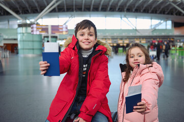 Handsome preadolescent boy and cute little girl in warm parka jackets smiling toothy smile standing at international airport and holding travel passport documents with air tickets and boarding pass