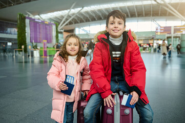 Cheerful Caucasian children- boy and girl in warm clothes, sitting on a suitcase and holding passports, with air tickets and boarding pass at the international airport. Family airplane travel concept