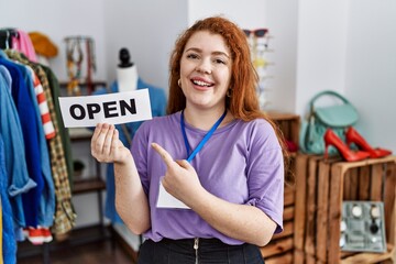 Young redhead woman holding banner with open text at retail shop smiling happy pointing with hand and finger