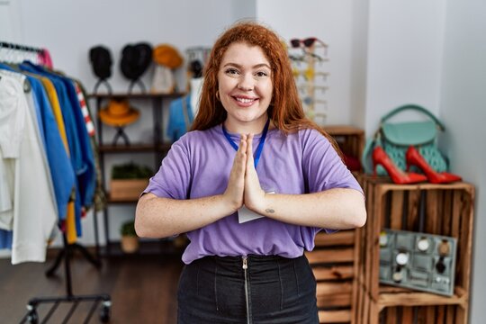 Young redhead woman working as manager at retail boutique praying with hands together asking for forgiveness smiling confident.