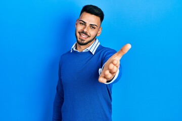 Fototapeta premium Young hispanic man with beard wearing casual blue sweater smiling cheerful offering palm hand giving assistance and acceptance.