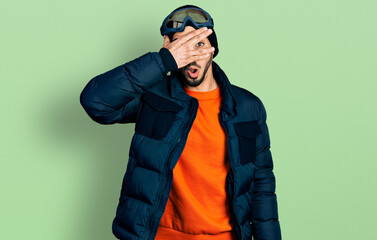 Young hispanic man with beard wearing snow wear and sky glasses peeking in shock covering face and eyes with hand, looking through fingers with embarrassed expression.