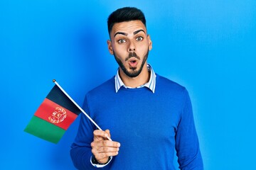 Young hispanic man with beard holding afghanistan flag scared and amazed with open mouth for surprise, disbelief face