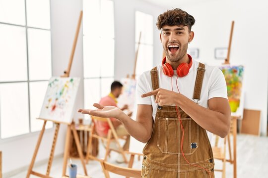 Young hispanic man at art studio amazed and smiling to the camera while presenting with hand and pointing with finger.