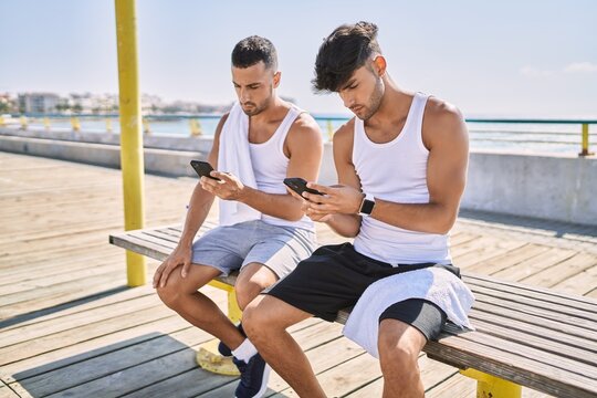 Two hispanic men sporty couple using smartphone sitting on bench at seaside