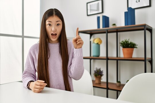 Young chinese girl wearing casual clothes sitting on the table at home pointing finger up with successful idea. exited and happy. number one.