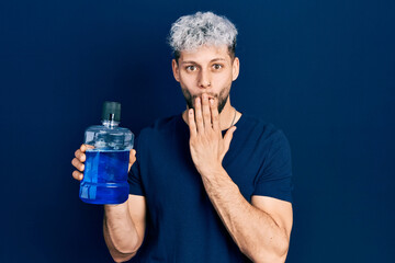 Young hispanic man with modern dyed hair holding mouthwash for fresh breath covering mouth with...