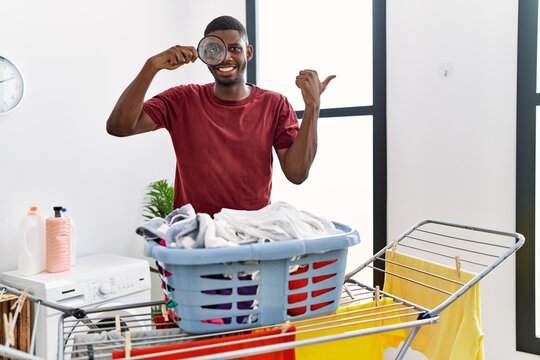 Young african american man holding magnifying glass looking for stain at clothes pointing thumb up to the side smiling happy with open mouth