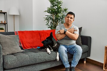 Young latin man and dog sitting on the sofa at home pointing to both sides with fingers, different direction disagree