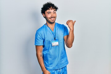 Young hispanic man wearing blue male nurse uniform smiling with happy face looking and pointing to the side with thumb up.