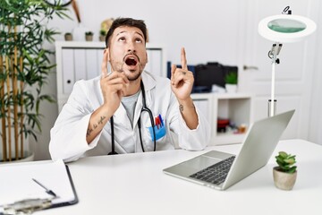 Young doctor working at the clinic using computer laptop amazed and surprised looking up and pointing with fingers and raised arms.