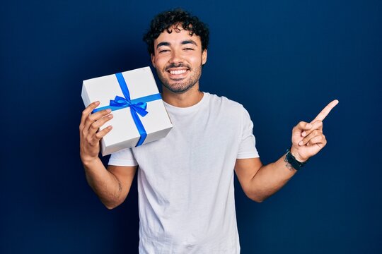 Young hispanic man holding gift smiling happy pointing with hand and finger to the side