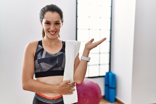 Young brunette woman wearing sportswear and towel at the gym smiling cheerful presenting and pointing with palm of hand looking at the camera.