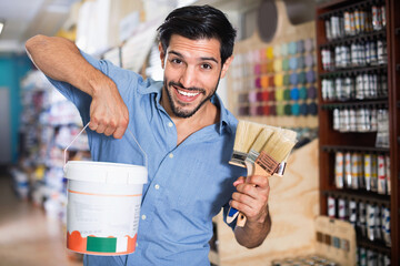 Satisfied young cheerful positive smiling man standing amongst racks in paint store with brushes...