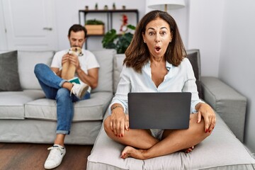 Hispanic middle age couple at home, woman using laptop afraid and shocked with surprise expression, fear and excited face.
