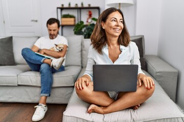 Hispanic middle age couple at home, woman using laptop looking away to side with smile on face, natural expression. laughing confident.