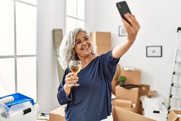 Middle age grey-haired woman toasting with wine making selfie by the smartphone at new home.