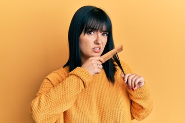Young hispanic woman styling hair using comb clueless and confused expression. doubt concept.