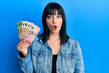 Young hispanic woman holding argentine pesos banknotes scared and amazed with open mouth for...