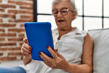 Senior grey-haired woman using touchpad sitting on sofa at home