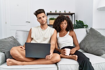 Fototapeta premium Young interracial couple using laptop at home sitting on the sofa looking sleepy and tired, exhausted for fatigue and hangover, lazy eyes in the morning.