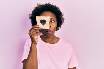 Young african american woman holding bread loaf with heart shape making fish face with mouth and squinting eyes, crazy and comical.