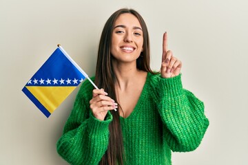 Young hispanic girl holding bosnia herzegovina flag smiling with an idea or question pointing finger with happy face, number one - obrazy, fototapety, plakaty