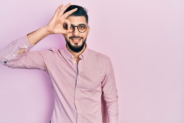 Handsome man with beard wearing casual shirt and glasses smiling happy doing ok sign with hand on eye looking through fingers