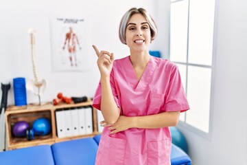 Young nurse woman working at pain recovery clinic smiling happy pointing with hand and finger to the side