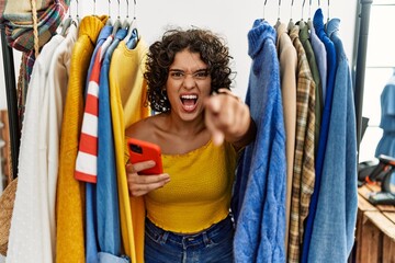 Young hispanic woman searching clothes on clothing rack using smartphone pointing displeased and frustrated to the camera, angry and furious with you