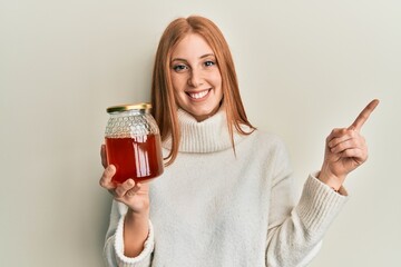 Young irish woman holding jar with honey smiling happy pointing with hand and finger to the side