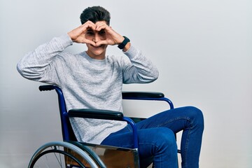 Young hispanic man sitting on wheelchair doing heart shape with hand and fingers smiling looking through sign