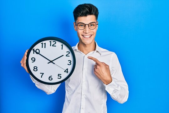 Young hispanic man holding big clock smiling happy pointing with hand and finger