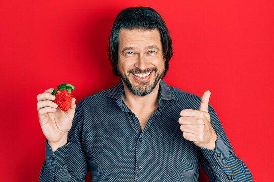 Middle age caucasian man holding strawberry smiling happy and positive, thumb up doing excellent and approval sign