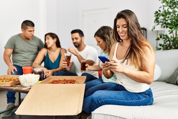 Group of young hispanic friends eating italian pizza sitting on the sofa. Woman smiling happy and using smartphone at home.