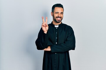 Handsome hispanic man with beard wearing catholic priest robe smiling with happy face winking at...