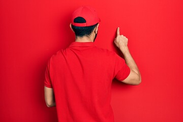 Hispanic man with beard wearing delivery uniform and cap posing backwards pointing ahead with finger hand