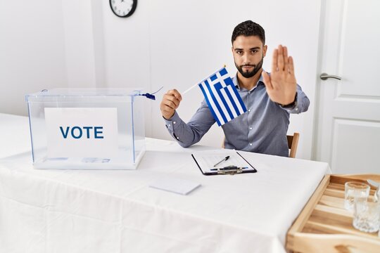 Young handsome man with beard at political campaign election holding greece flag with open hand doing stop sign with serious and confident expression, defense gesture