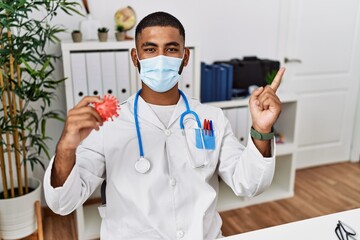 Fototapeta premium Young indian doctor holding virus toy wearing safety mask smiling happy pointing with hand and finger to the side