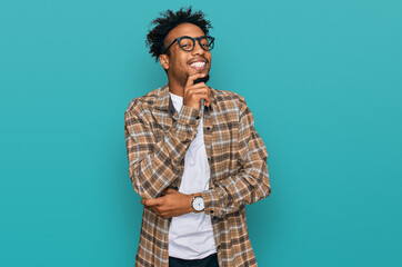 Young african american man with beard wearing casual clothes and glasses looking confident at the camera smiling with crossed arms and hand raised on chin. thinking positive.